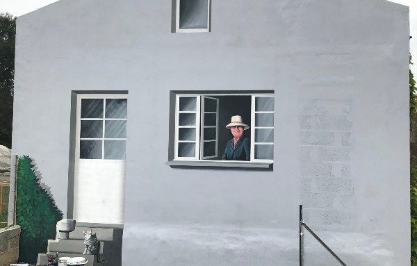 fresque trompe oeil Homme portant un chapeau, regardant par une fenêtre d'une maison grise.
